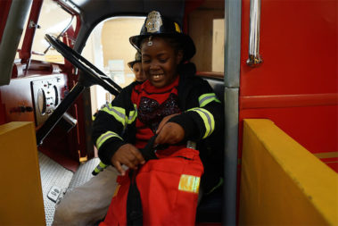 Child wearing firefighter gear within a fire truck.