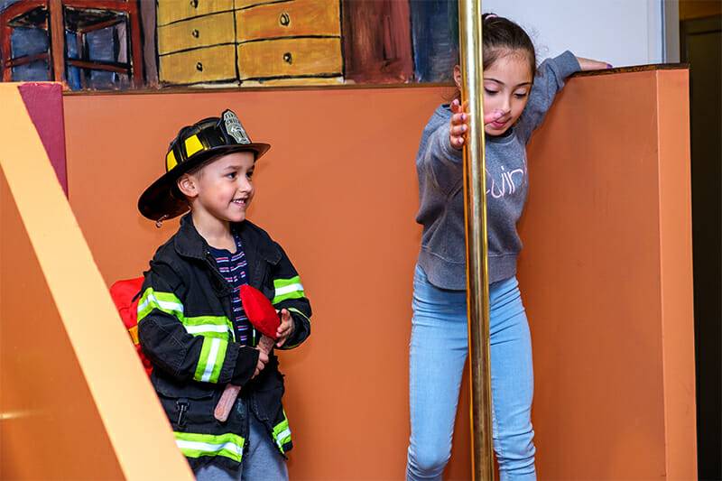 Two children playing within our firefighter exhibit, one dressed in firefighter gear, the other sliding down a pole.