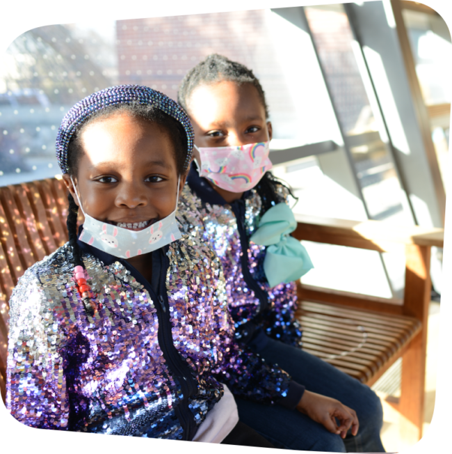 two girls on bench girl with mask down smiling next to girl with mask on sitting on bench with sun shining behind them through a window