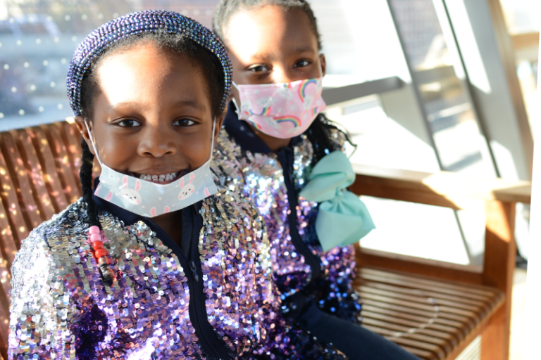 girl with mask down smiling next to girl with mask on sitting on bench with sun shining behind them through a window