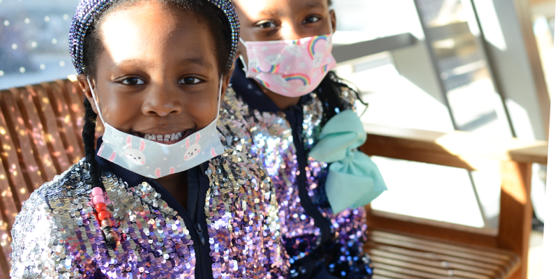 girl with mask down smiling next to girl with mask on sitting on bench with sun shining behind them through a window