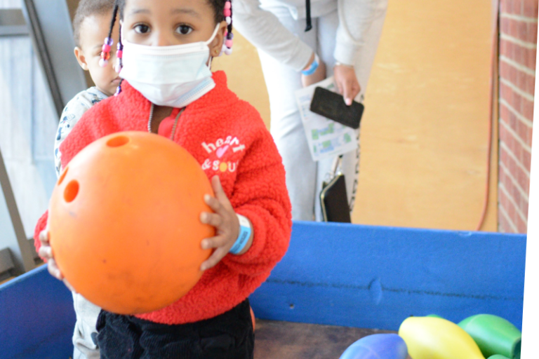 Young child holding a large orange bowling ball in the bowling lane with several colorful pins knocked down behind her