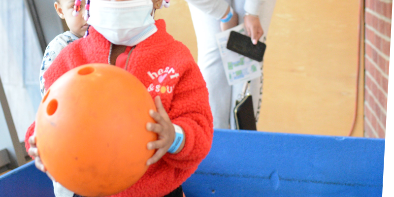 Young child holding a large orange bowling ball in the bowling lane with several colorful pins knocked down behind her
