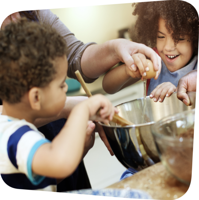 children with curly hair and light brown skin gleefully helping an adult add ingredients to a bowl for mixing
