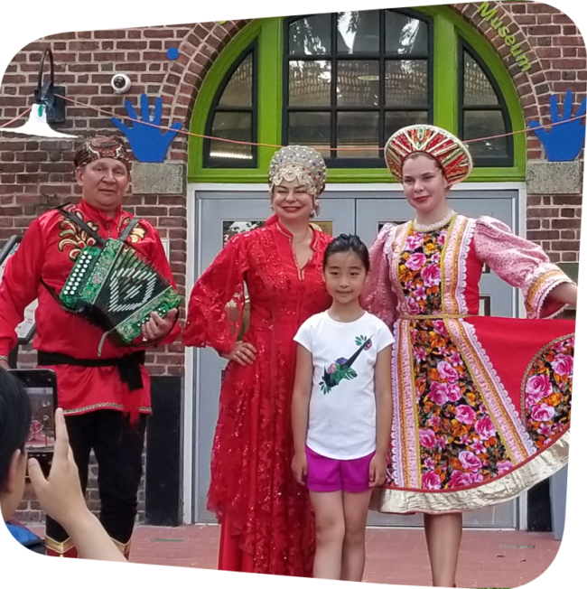 Performers wearing cultural attire posing for a picture with a smiling child in front of the children's museum.
