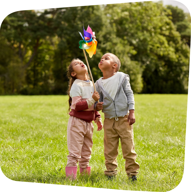 Two children blowing on a pinwheel in the middle of a meadow with evergreen trees in the background