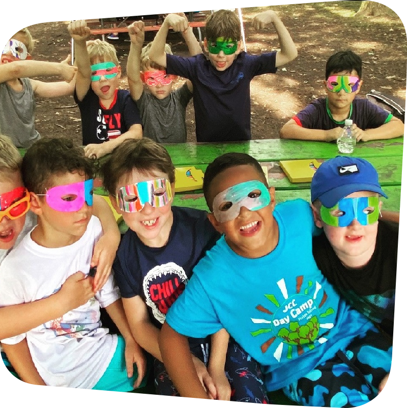 children in decorated eye masks leaning against each other sitting on a picnic bench