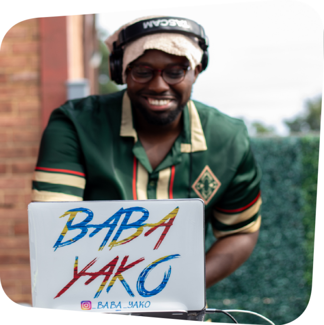 Smiling DJ Baba Yako standing behind a white laptop that displays his name in blue, red, and yellow letters. DJ Baba Yako wears headphones on his head and a green shirt.