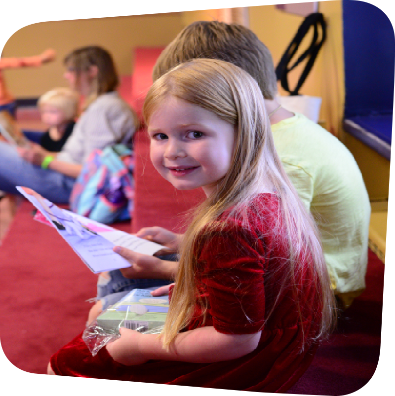 Central foreground: Two children attending the LINC storytime reading event. One of the children, a girl, smiles at the camera. Beside her, hidden by her frame, sits a boy reading a book.