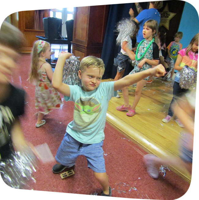 Inside a room, a young boy in shorts and a t-shirt dances at the center of the photo, with more moving children surrounding.