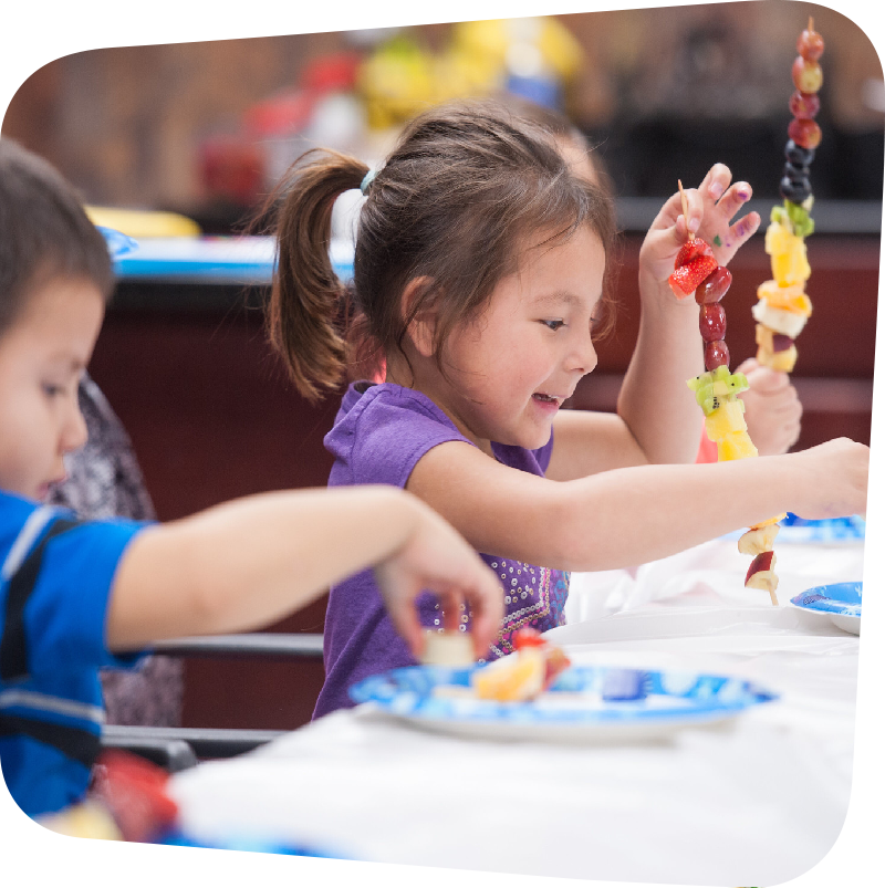 Two young children seated at table putting fruit and stick skewers. Smiling seated girl is central focus of image.