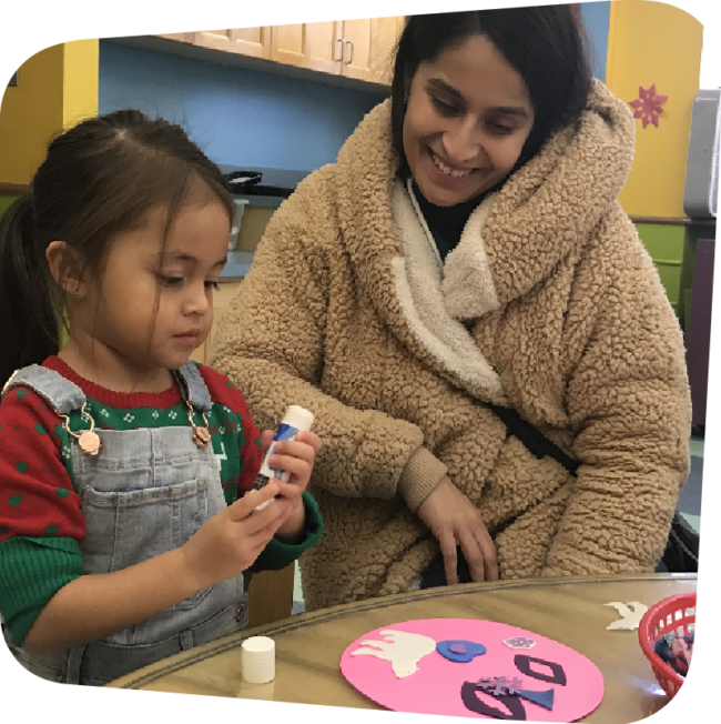 A mother in a brown teddy-bear textured jacket smiles down at her daughter as the little girl focuses on twisting a glue stick. On the table they sit in front of is a pink circle decorated with white, blue, and purple cut-outs.