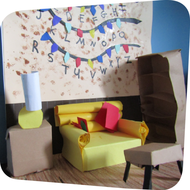 up-close of a mini model of the Walk-in Workshop craft. On a folded piece of paper, a multi-colored paper decoration hung on the wall with the letters of the alphabet. In front of the wall, a living room area with a table and lamp, beside which is a yellow couch, beside which is a brown bookcase, and a coffee table in front of the couch. These objects were folded to make them three-dimensional. The paper floor of model was decorated with a brown wood pattern.