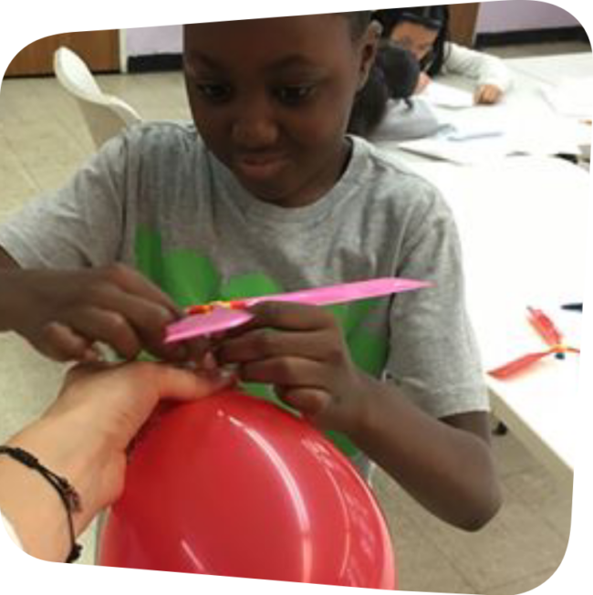 smiling boy holding a red balloon attaching a propeller to it. photo courtesy of Crazy Science