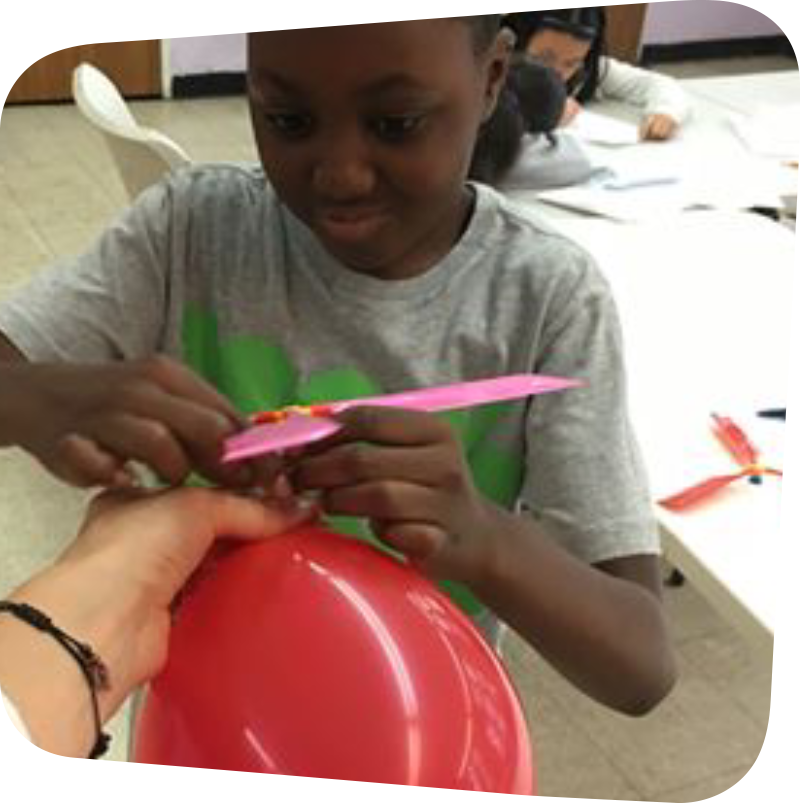 smiling boy holding a red balloon attaching a propeller to it. photo courtesy of Crazy Science