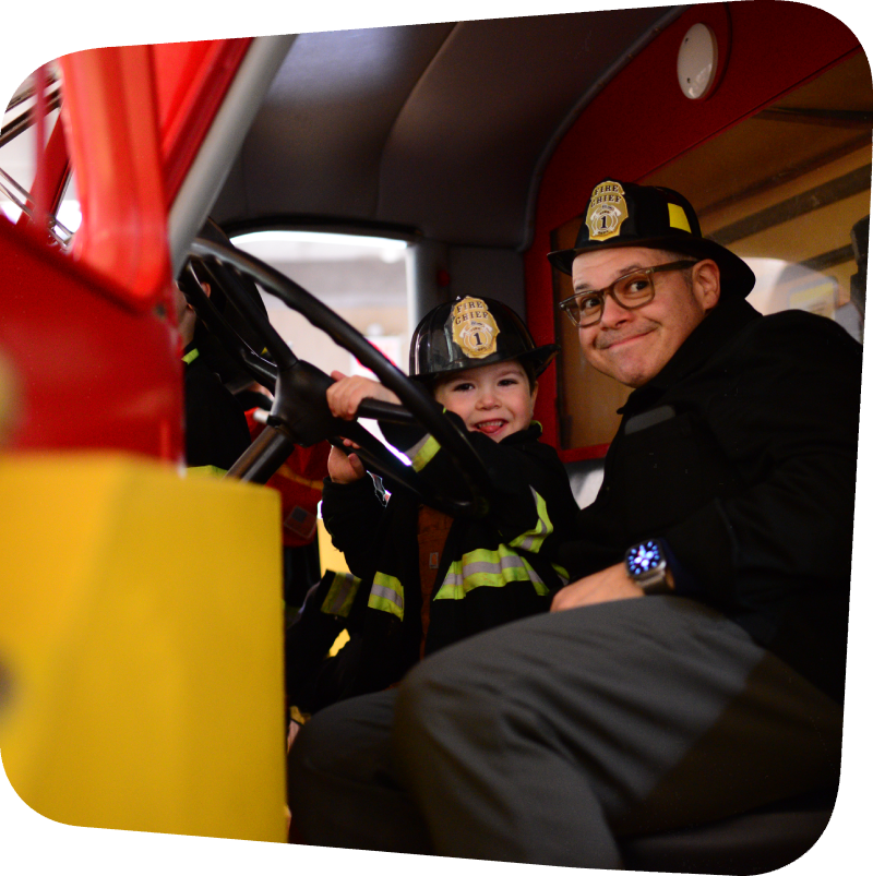 a man and child dressed in firefighter costumes in the Ladder 11 fire engine at the Staten Island Children's Museum
