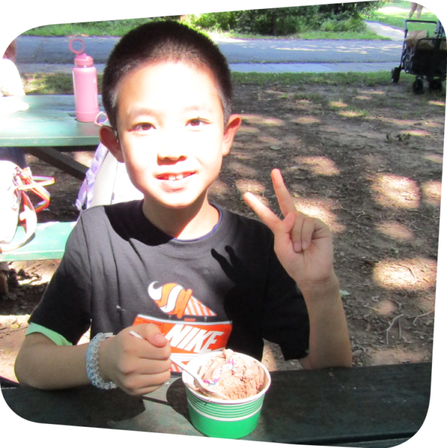 smiling boy holding up two fingers with a cup of ice cream in front of him