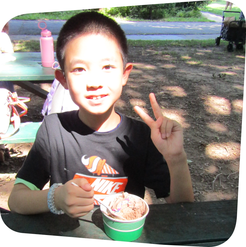 smiling boy holding up two fingers with a cup of ice cream in front of him