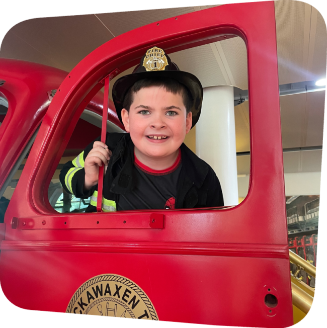 smiling boy wearing a firefighters outfit standing in the door of ladder 11 exhibit at The Staten Island Children's Museum