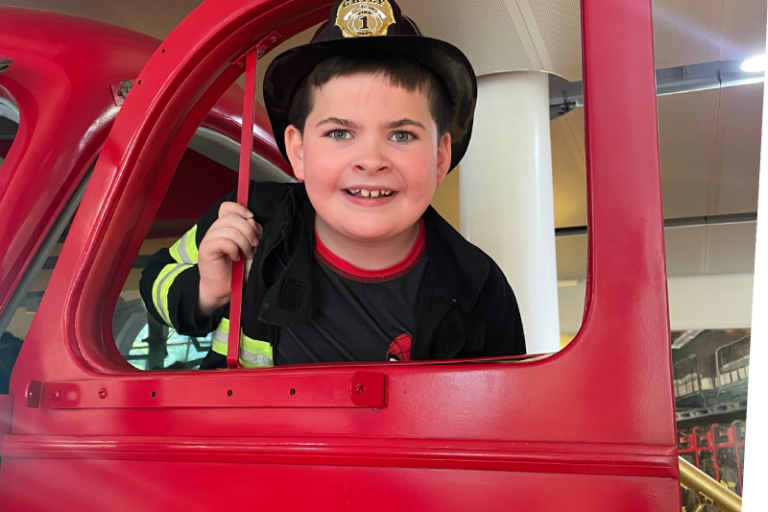 smiling boy wearing a firefighters outfit standing in the door of ladder 11 exhibit at The Staten Island Children's Museum