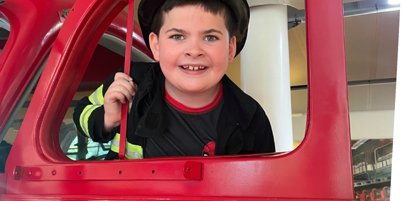 smiling boy wearing a firefighters outfit standing in the door of ladder 11 exhibit at The Staten Island Children's Museum